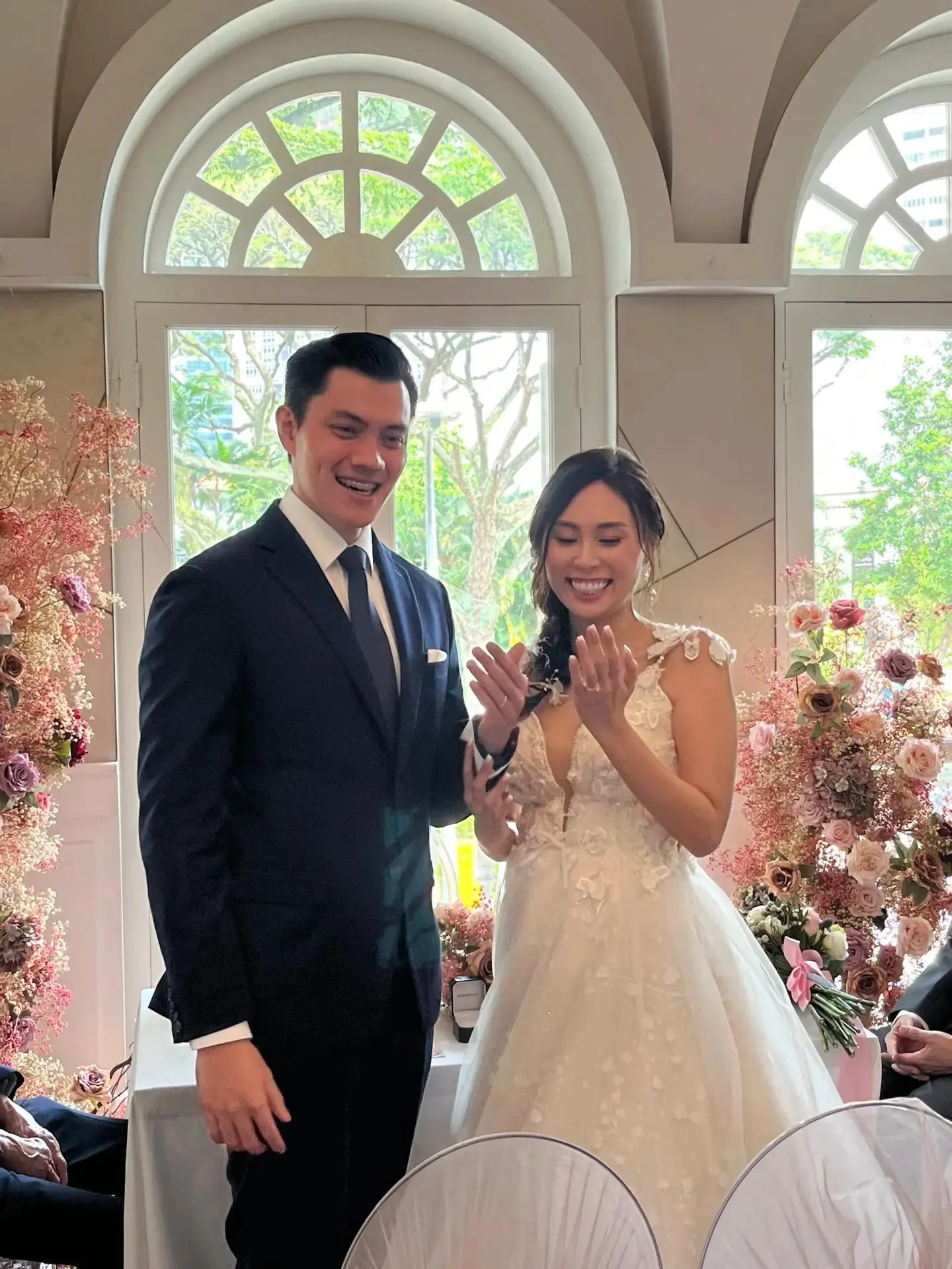 Jia En and Charlotte beaming as they admire their new wedding rings during their solemnisation at The Secret Patio, framed by arched windows and pink floral arrangements