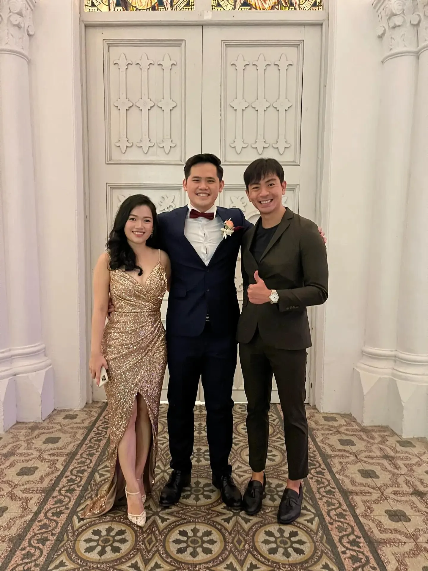 The emcee smiling with the groom and a guest in evening wear in front of the white chapel doors at CHIJMES