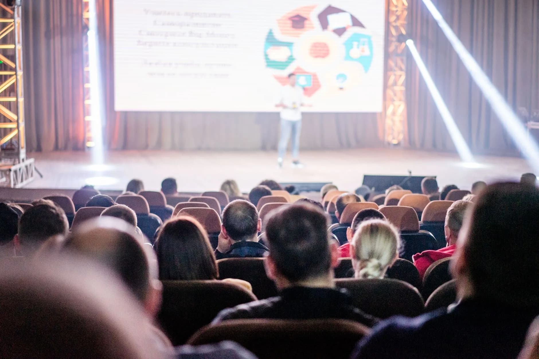 Presenter on stage with a large screen and spotlights before a seated audience
